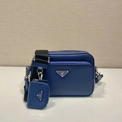 A small blue leather camera bag with a wide webbing strap, shown beside its matching zip pouch