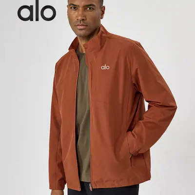 A man wearing an unzipped rust brown shell jacket over an olive top, hands in pockets, against a pale studio wall